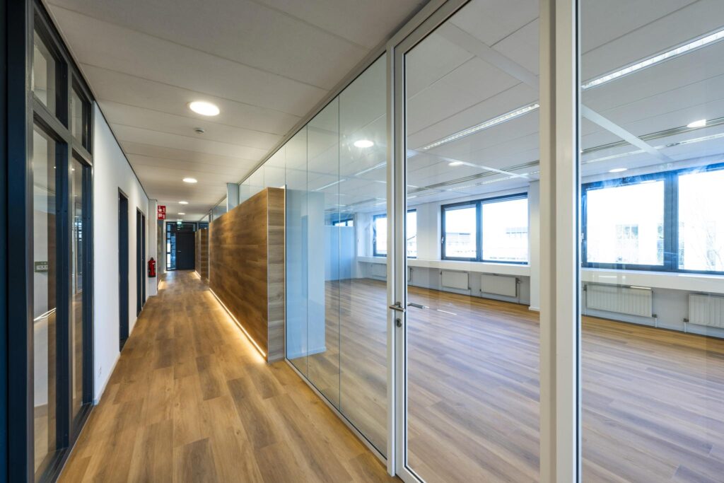 Modern office hallway with wood flooring, glass walls, and a view into a bright, empty workspace.